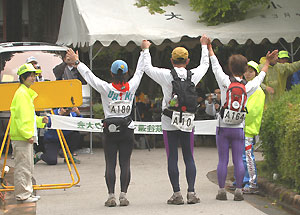 2004年、10回目の連続完踏する時の写真（両側の女性と3人が手を取り合って万歳しながらゴールテープを切る瞬間。左はＵＭＭＬのＹ子さん、右は京都・鞍馬のＭさんの奥様）（ゴールテープを持つボランティアさんと、正面には大会本部のテントが写っています）