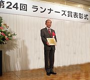 授賞式：壇上で盾を持つ筆者の写真（胸に赤いハンカチ、赤いネクタイ、後ろには「第２４回ランナーズ賞表彰式」の看板）